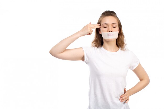 A front view young female in white t-shirt with white bandage around her mouth pointing fingers into her head on the white