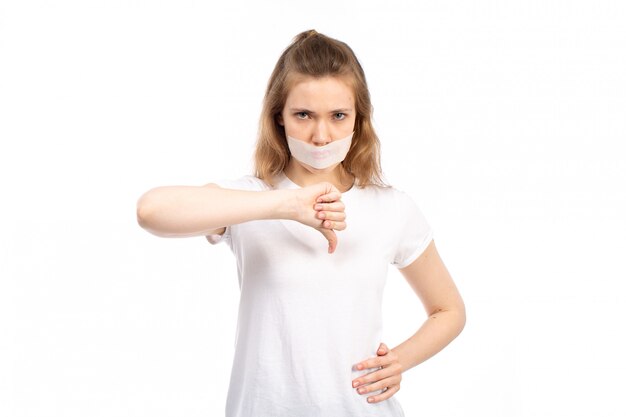 A front view young female in white t-shirt with white bandage around her mouth showing unlike sign on the white