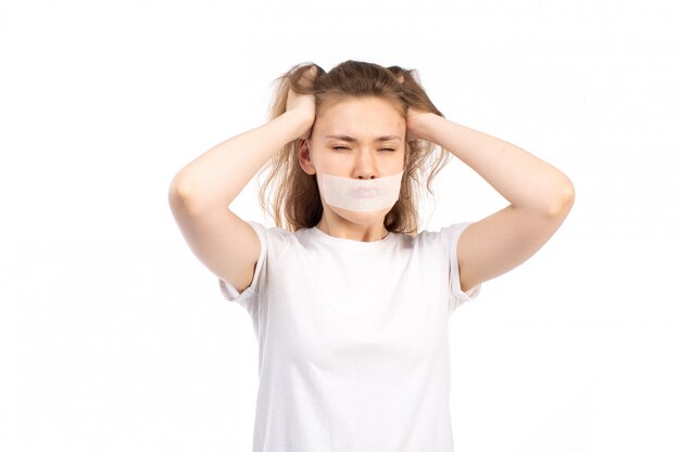 A front view young female in white t-shirt with white bandage around her mouth touching her hair angry on the white