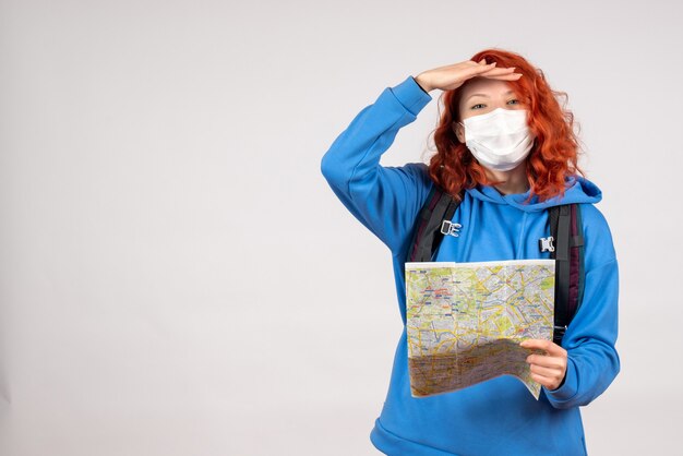 Front view of young female with backpack and map in mask on white wall