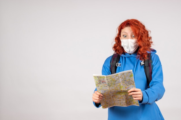 Front view of young female with backpack and map in mask on white wall
