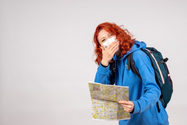 Front view of young female with backpack and map in mask on white wall