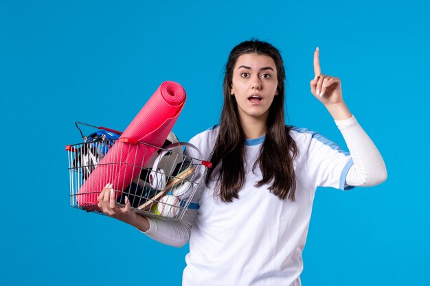 Вид спереди молодая женщина с корзиной, полной спортивных вещей, синяя стена