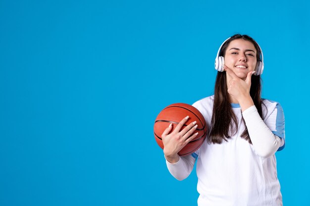 Вид спереди молодая женщина с наушниками, держащая баскетбол на синей стене