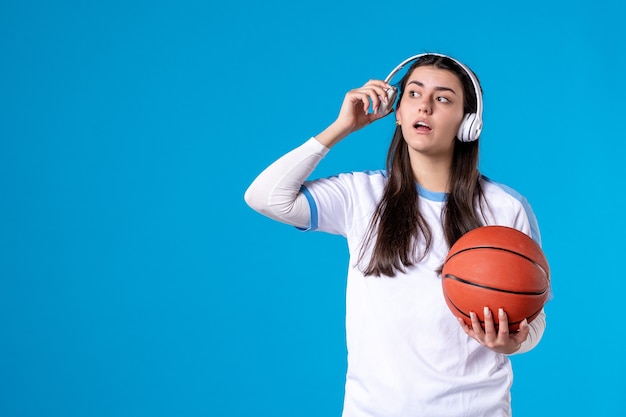 青い壁にバスケットボールを保持しているイヤホンを持つ若い女性の正面図