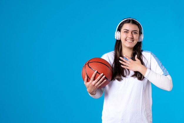 Вид спереди молодая женщина с наушниками, держащая баскетбол на синей стене