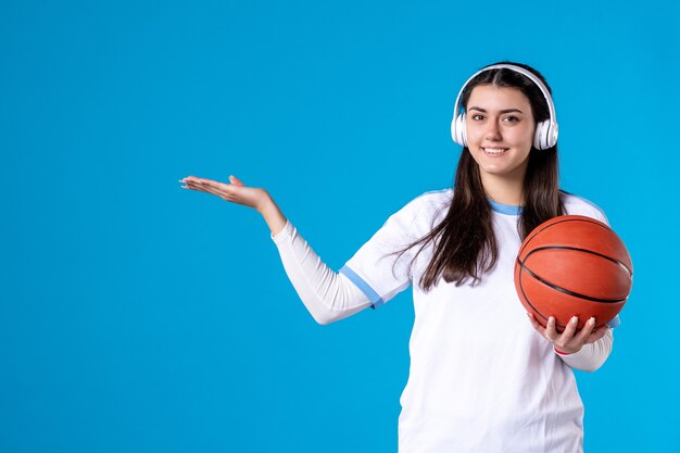 青い壁にバスケットボールを保持しているイヤホンを持つ若い女性の正面図