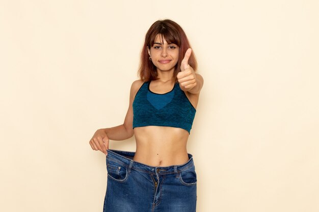 Front view of young female with fit body in blue shirt and jeans smiling on light-white wall