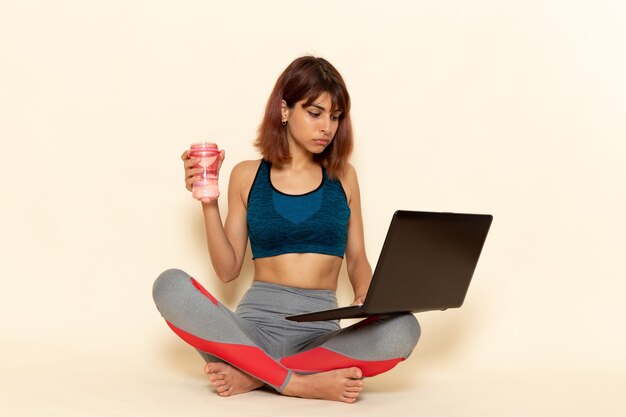 Front view of young female with fit body in blue shirt using laptop on white wall