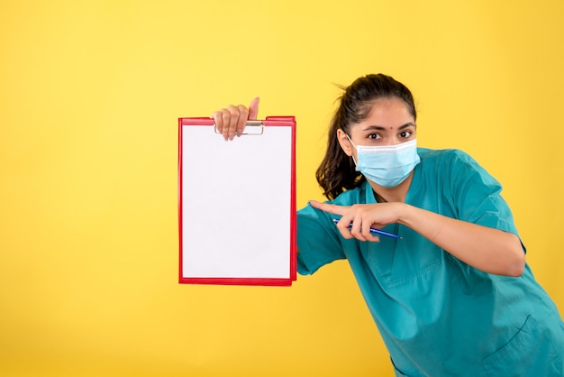 Front view of young female with medical mask pointing at clipboard on yellow wall