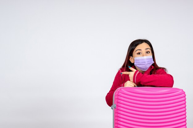 白い壁のウイルスの女性の休暇のcovidカラーパンデミック旅行のマスクでピンクのバッグを持つ若い女性の正面図