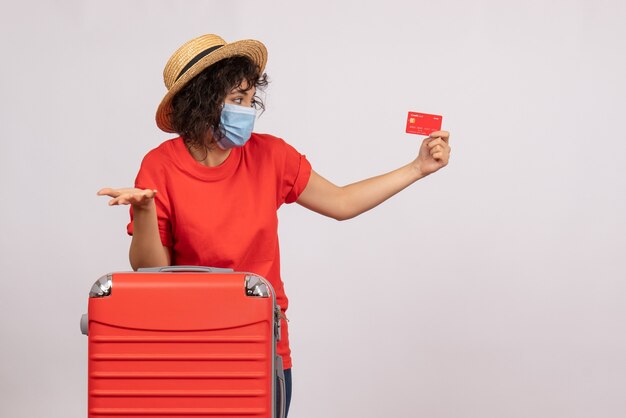 Front view young female with red bag in mask holding bank card on white background sun covid pandemic vacation trip tourist colors