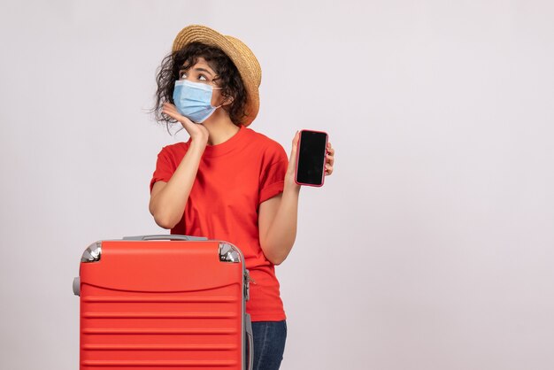 Вид спереди молодая женщина с красной сумкой в маске, держащая телефон на белом фоне, солнце, пандемия, отпуск, туристический вирус, цвет