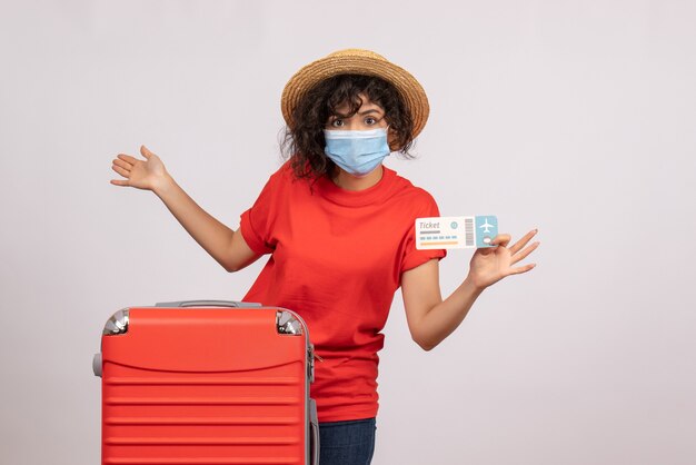 Вид спереди молодая женщина с красной сумкой в маске, держащая билет на белом фоне, солнце, пандемия, отпуск, туристический цвет