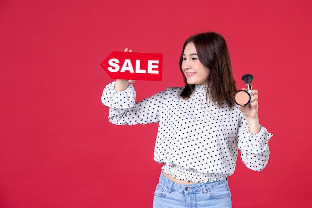 Free photo front view young female with tassel powder for make-up and sale nameplate on red wall