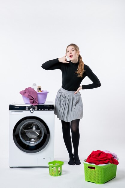 Вид спереди молодой женщины со стиральной машиной и грязной одеждой на белой стене