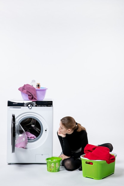 Вид спереди молодой женщины со стиральной машиной, складывающей грязную одежду на белой стене