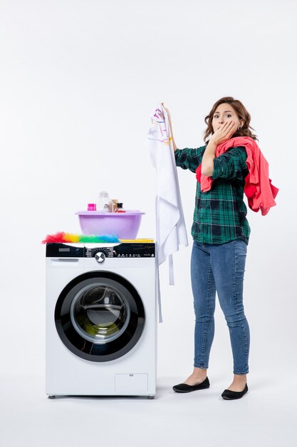 Front view of young female with washing machine preparing clothes for wash on the white wall