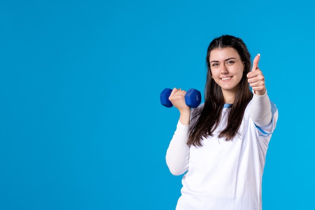 Front view young female working out with blue dumbbells on blue wall