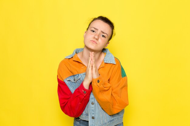 A front view young female in yellow shirt colorful jacket and blue jeans posing and praying