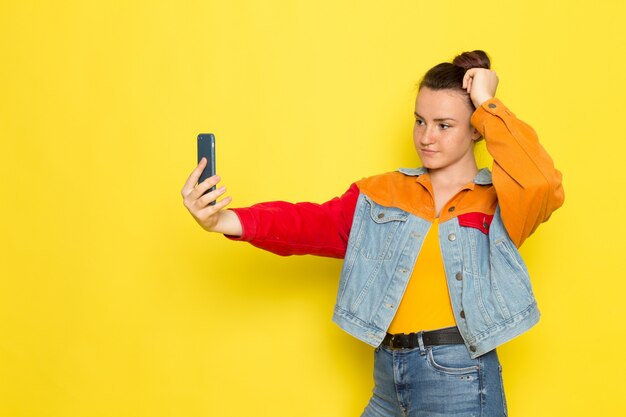 A front view young female in yellow shirt colorful jacket and blue jeans posing and taking a selfie