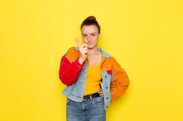 A front view young female in yellow shirt colorful jacket and blue jeans posing and threatening