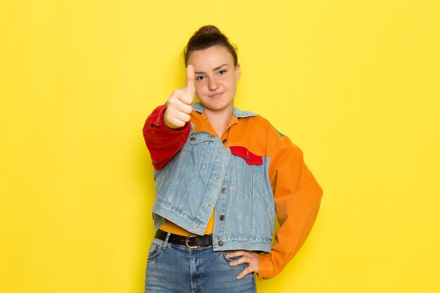 黄色のシャツのカラフルなジャケットとブルージーンズのポーズで正面の若い女性