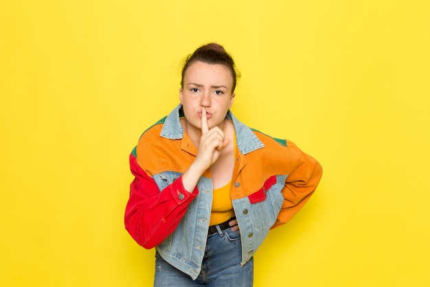 A front view young female in yellow shirt colorful jacket and blue jeans showing silence sign