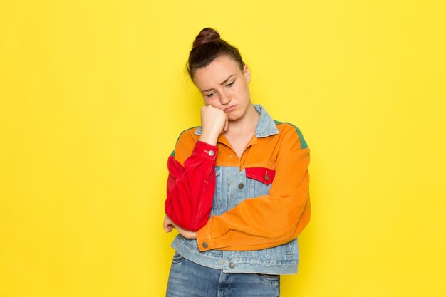 A front view young female in yellow shirt colorful jacket and blue jeans with thinking expression