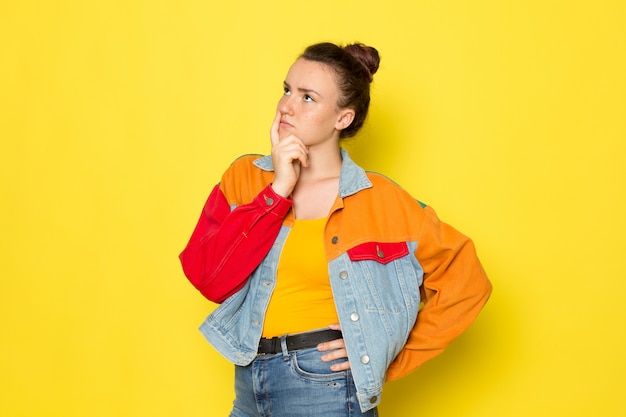 A front view young female in yellow shirt colorful jacket and blue jeans with thinking expression