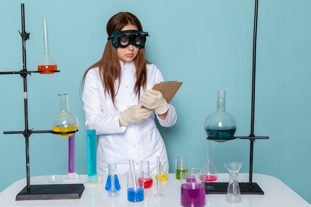 Front view of young feman chemist in white suit in front of table writing down notes