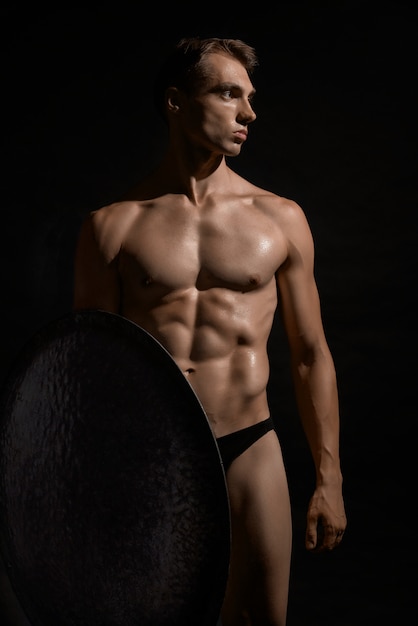 Front view of young fit strong warrior with perfect muscular body. Tensed athlete in underwear looking away, posing and holding shield on black  wall. Cocept of warrior, strength.