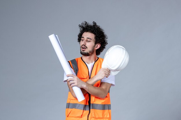 Front view of young focused male constructor in warning vest holding safety helmet and showing blank on gray wall