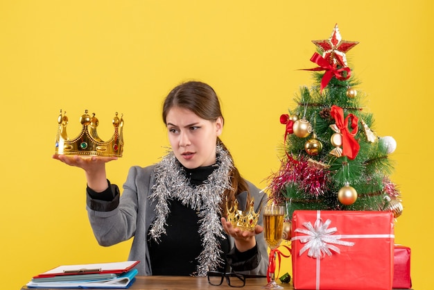 Вид спереди молодая девушка держит большую и маленькую корону и проверяет большую рождественскую елку и подарочный коктейль