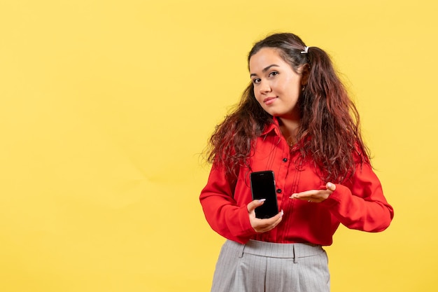 Front view young girl in red blouse with cute hair holding her phone on a yellow background child color kid girl youth innocence