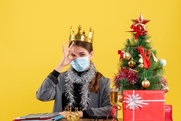 Молодая девушка с медицинской маской, вид спереди, делает знак оки перед ее глазом, рождественская елка и подарочный коктейль