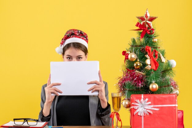 Front view young girl with xmas hat sitting at the table covering her face with documents xmas tree and gifts cocktail