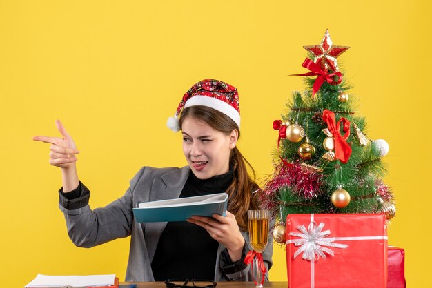 Front view young girl with xmas hat sitting at the table taking document folder xmas tree and gifts cocktail