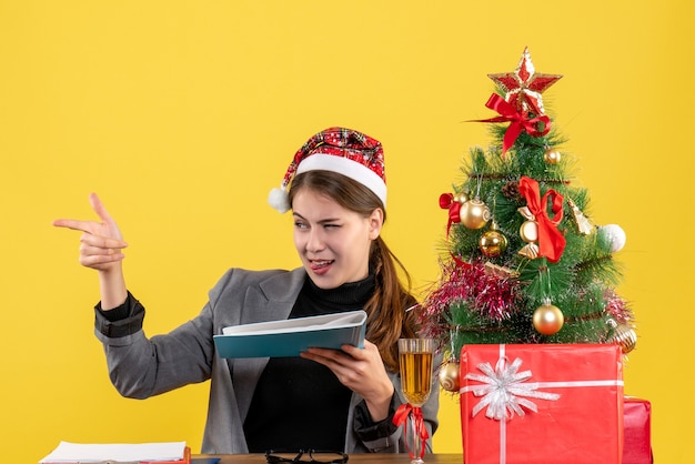 Молодая девушка с рождественской шляпой сидит за столом, принимая папку с документами, рождественскую елку и подарки, коктейль, вид спереди