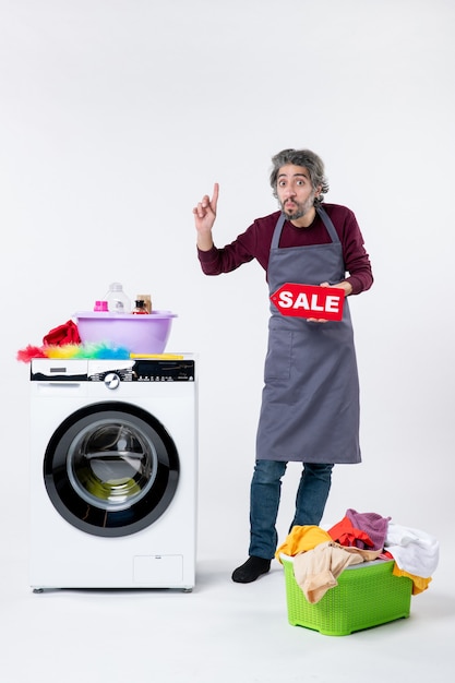 Front view young guy in apron holding up sale sign pointing with finger up near washer on white wall