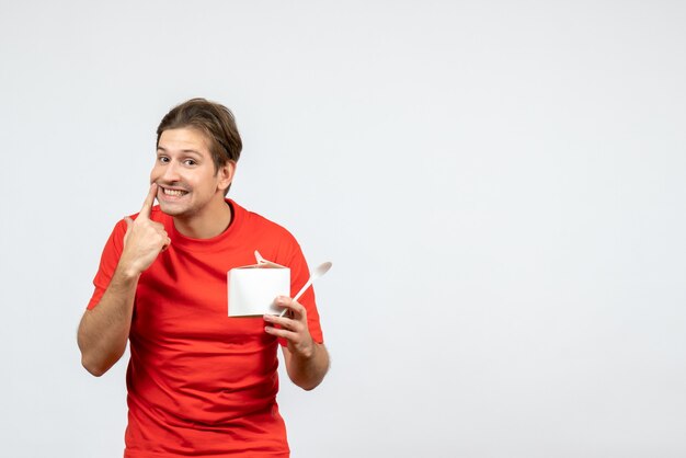Вид спереди молодого парня в красной блузке, держащего бумажную коробку и ложку, делая жест на белом фоне