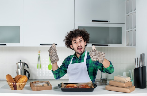 Front view of young guy wearing holder standing behind table freshly-baked pastry on it making ok gesture in the white kitchen