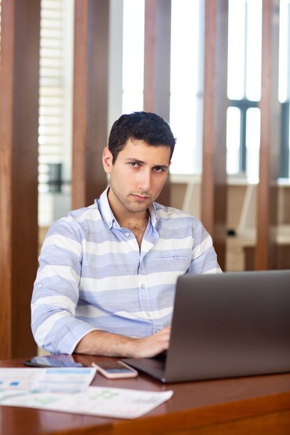A front view young handsome man in striped shirt working inside conference hall using his silver laptop during daytime work activity building