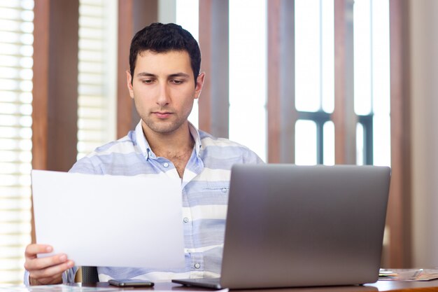 A front view young handsome man in striped shirt working inside conference hall using his silver laptop looking through documents during daytime work activity building