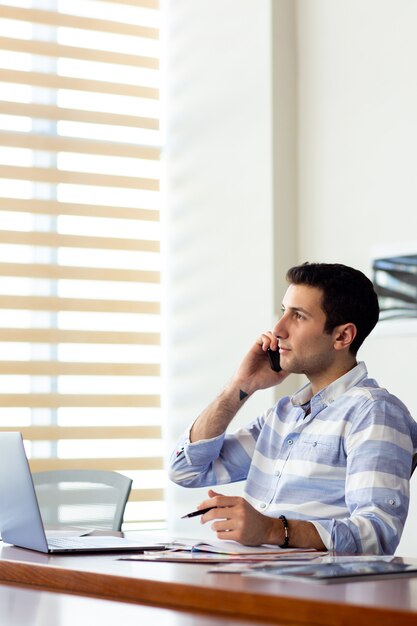 A front view young handsome man in striped shirt working inside conference hall using his silver laptop looking through documents talking on the phone during daytime work activity building