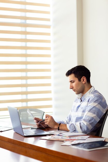 A front view young handsome man in striped shirt working inside conference hall using his silver laptop looking through documents writing down during daytime work activity building