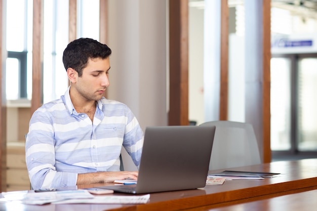주간 작업 활동 건물 동안 그의 은색 노트북을 사용하여 자신의 사무실에서 일하는 스트라이프 셔츠에 전면보기 잘 생긴 젊은이