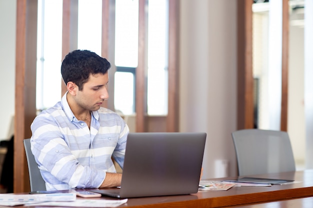 Un giovane uomo bello di vista frontale in camicia a strisce che lavora dentro il suo ufficio facendo uso del suo computer portatile d'argento durante la costruzione di attività di lavoro di giorno