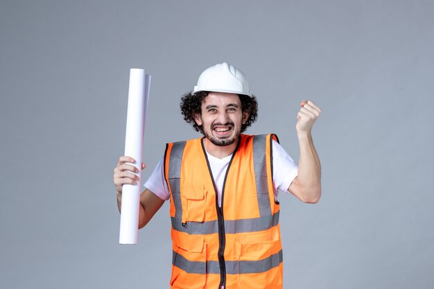 Front view of young happy construction worker in warning vest with safety helmet and showing blank on gray wall