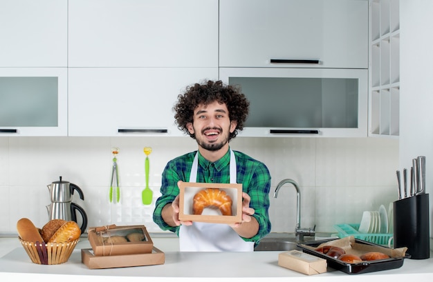 Foto gratuita vista frontale del giovane uomo felice che mostra la pasticceria appena sfornata in una piccola scatola nella cucina bianca